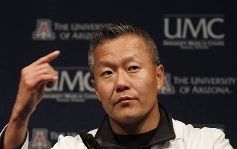 Peter M. Rhee talking on stage and pointing at something while wearing a white doctors attire with a black shirt underneath.