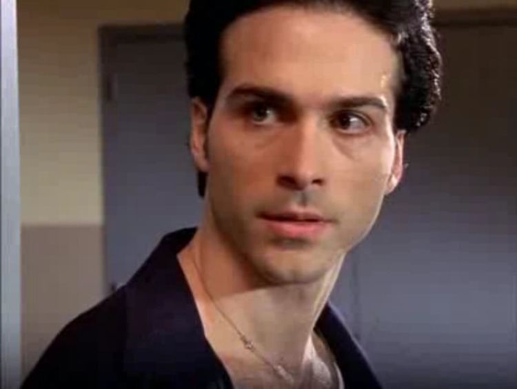 Peter Schibetta wearing dark blue long sleeves and a necklace in a scene from the 1997 tv series Oz