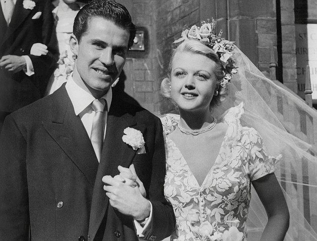 Peter Shaw with Angela Lansbury both smiling and holding each other's hands after their wedding day at the Chapel of St. Columba's Church House in Lennox Gardens, Kensington, London, 12th August 1949; with a couple in the background. Peter is wearing a white collared shirt and a necktie  under a black coat with a corsage on his left chest while Angela is wearing a pearl layered necklace, hoop earrings, a flower headdress and a veil, while on a deep-V collared flower-patterned wedding dress.