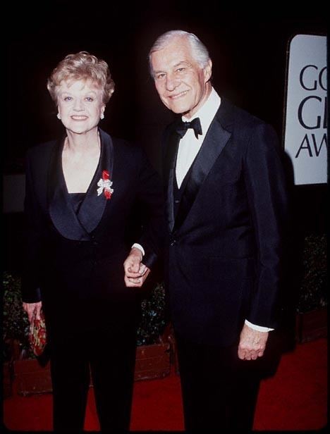 Peter Shaw and his wife, Angela Lansbury both smiling, holding hands while standing on a red carpet during the Governor's Global Award with its banner in the background. Angela with a curly blonde hair, holding a red patterned purse in her right hand, wearing a pair of earrings in a pair of black slacks, a red and white corsage pinned on her left chest over a black long sleeve coat with a black inner tube; while Peter with white hair is wearing a pair of black slacks, a black bowtie over a white collared shirt and a black long sleeve coat.