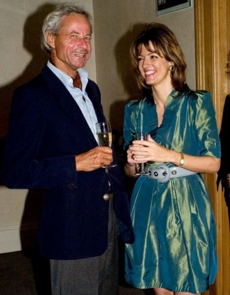 from left, Johannes Muehlenburg is smiling, standing, holding a champagne glass with his right hand, has gray hair, wearing a light blue polo under a black coat and a black pants, at the right, Petronella Wyatt is smiling, standing, holding a champagne glass with her hands, has long brown hair wearing hoop earrings, gold watch and a shiny blue green dress with white belt.