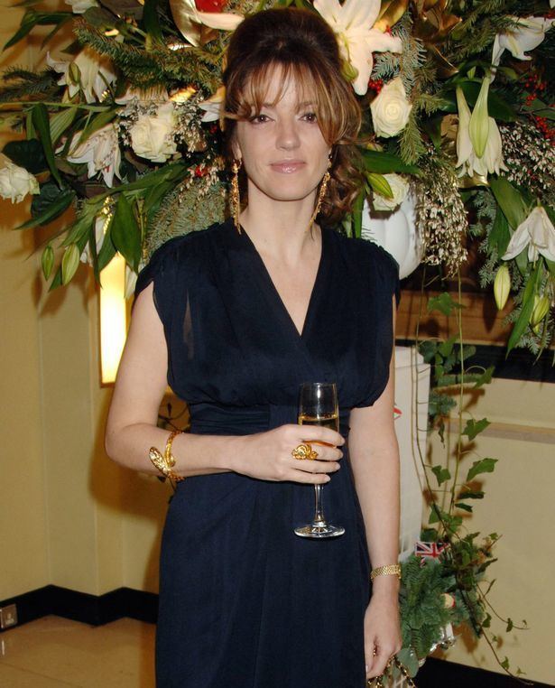 Petronella Wyatt is smiling, standing in a room with a large plant of white flowers and a green vine leaf hanging at the back, holding a champagne glass, has brown hair wearing gold chandelier earrings, a gold bracelet, a ring and a black long dress.