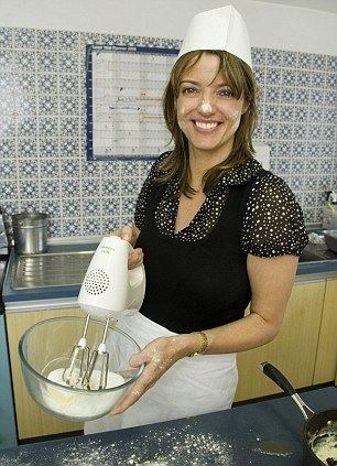 Petronella Wyatt is smiling, baking, while holding a white mixer with her right hand and holding a bowl with her left hand, standing in the middle of the kitchen with blue printed tiles walls, brown cabinets, gray countertops and a stainless steel sink, she has long brown hair, flour on her cheek and nose, wearing a white chef toque, black with dotted sleeve, in front is a gray countertop with flour dusted on top and a black frying pan on the bottom right.