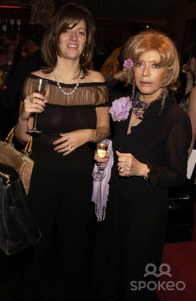 In a party at the right,  Petronella Wyatt is smiling, standing, holding a champagne glass with her right hand, a brown bag on her right elbow and left arm bend over her stomach, has brown hair wearing a white beads necklace a hoop earrings, silver ring on left and right hand, gold watch, and a black long dress, at the right, Veronica Banszky Von Ambroz is serious, standing while holding a champagne glass with her right hand, left hand closed, has blonde hair with a purple flower, wearing a long chandelier earrings, black bead necklace, silver ring, bracelet, and a black long dress with purple flower and purple scarf.