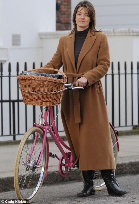 Petronella Wyatt is smiling, standing in the road, posing with her pink bike with brown basket in front, at the back is a black fence and a pathwalk, has brown hair wearing black top under a long brown coat with black boots.