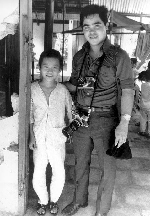 Young Phan Thi Kim Phuc smiling together with Nick Ut inside of a house with children in their background in an old photograph. Phan has black short hair wearing black sandals, a white long sleeve, and white pants while Nick’s hands are around Phan's shoulders. Nick has black hair and holding a black hat in his left hand wearing a watch on his left wrist, a shoulder bag, two cameras around his neck, black formal shoes, and a tuck-in black polo and black pants with a black belt