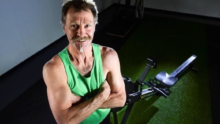 Phil Carman smiling with a mustache and crossed arms while standing beside his workout equipment