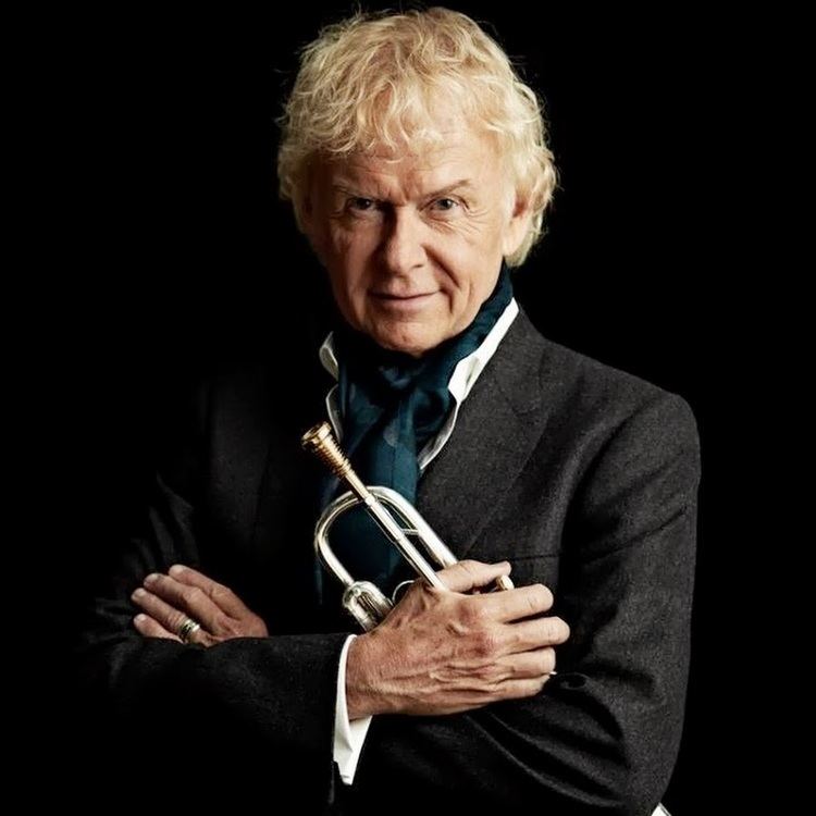Phil Driscoll smiling while holding a trumpet with blonde wavy hair and wearing a blue ribbon scarf, ring, and a white long sleeve under a black coat