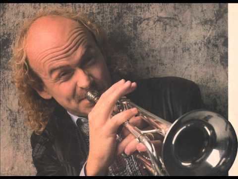 Phil Driscoll playing trumpet with a furrowed forehead and shoulder-length blonde curly hair while wearing a white long sleeve under a black and red cardigan and a black leather jacket