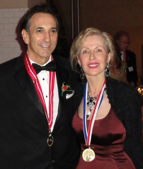 Philip Zepter smiling with the woman beside him and wearing a black coat, white long sleeves, black bow tie, and medallion
