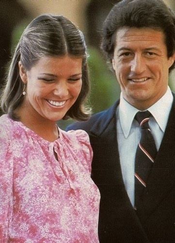 Philippe Junot smiling and wearing a black coat, white long sleeves, and necktie while Princess Caroline of Monaco wearing a pink dress