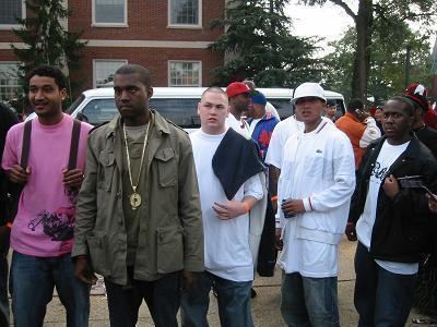 Kanye West (second from left), Plain Pat (center), and their colleagues are looking at something with serious faces. Kanye is wearing a greenish-grey jacket, medal, and black pants while Plain is wearing a white t-shirt, denim pants, and black cloth on his shoulder