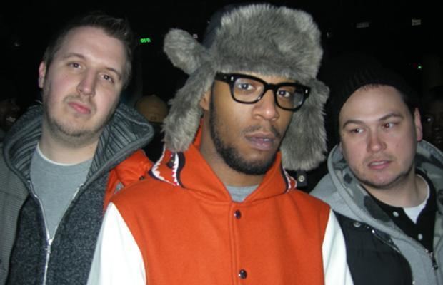 A man, Kid Cudi, and Plain Pat (left to right) with serious faces. The man on the left side is wearing a light gray shirt under a gray jacket. Kid is wearing a gray trapper hat, eyeglasses, gray shirt under a white and red jacket. Plain is wearing a black beanie and white shirt under a gray and black jacket