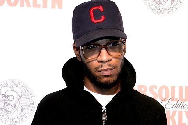 Kid Cudi with a sad face, mustache, and beard while wearing a black cap with letter C, eyeglasses, and white shirt under and black jacket