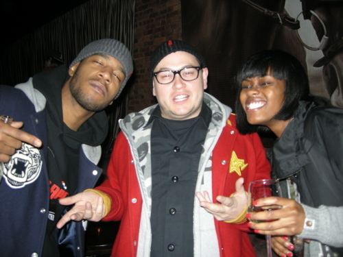 Kid Cudi, Plain Pat, and a woman (left to right) are smiling. Kid is wearing a gray beanie and black hoodie under a blue and gray jacket.  Plain is wearing a black beanie, eyeglasses, and a black shirt under a red and gray jacket. The woman on the right having a glass in hand is wearing a black and gray jacket.