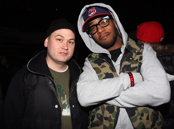 Plain Pat with a tight-lipped smile and Kid Cudi with a mustache, beard, and cross arms. Plain is wearing a black beanie and green shirt under a black jacket while Kid is wearing a blue and red cap, eyeglasses, black shirt under a light gray hoodie, and camouflage vest