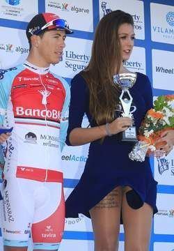 A podium girl holding a trophy and flowers with a cyclist beside him