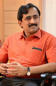 Ponraj Vellaichamy with a serious face while sitting on a steel chair, with a mustache, wearing an orange polo shirt, and gray pants.