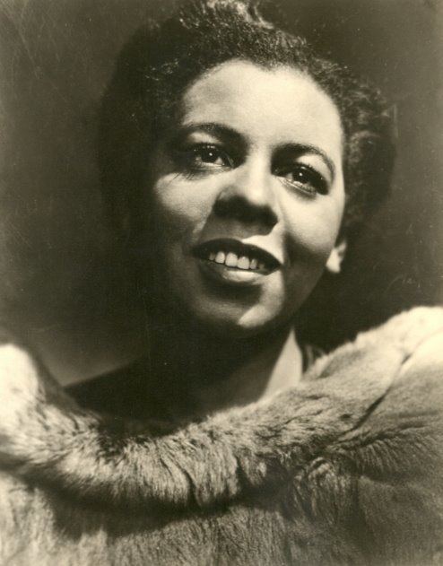 Portia White smiling while looking at something, with a tied-up hair, and wearing a blouse with fur