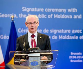 Herman Van Rompuy delivering a speech while using a podium and wearing eyeglasses and white long sleeve under a red necktie and black coat