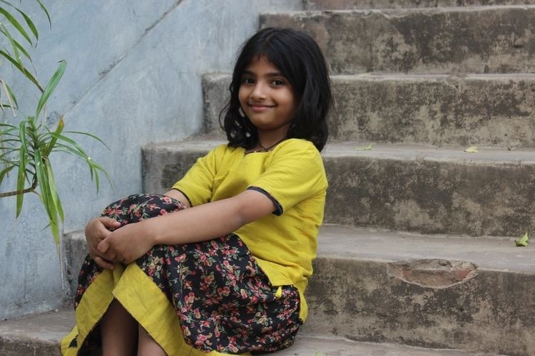A girl Potol from Potol Kumar Gaanwala wearing a yellow dress and sitting on the stairs