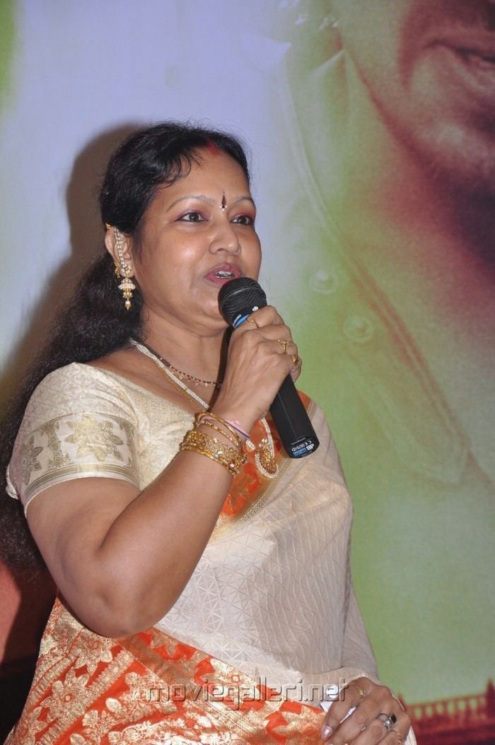 Prabha wearing earrings, a necklace, bracelets, rings, a yellow and orange dress while holding a microphone.