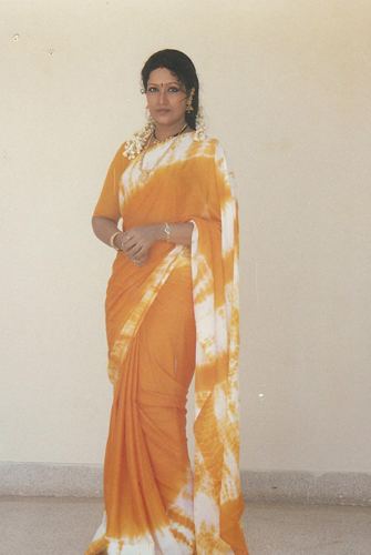 Prabha wearing earrings, a necklace, bracelets, and a yellow and white dress.