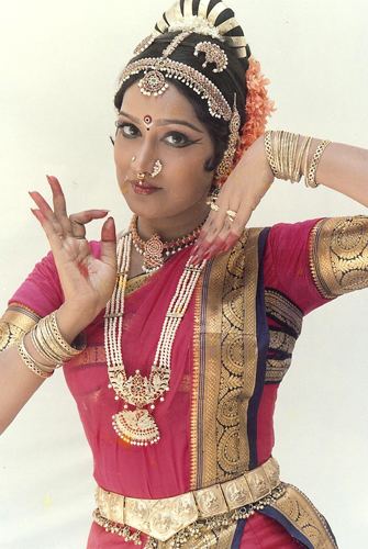 Prabha with nose piercings, wearing a headdress, earrings, a necklace, bracelets, rings, and a yellow and pink dress.