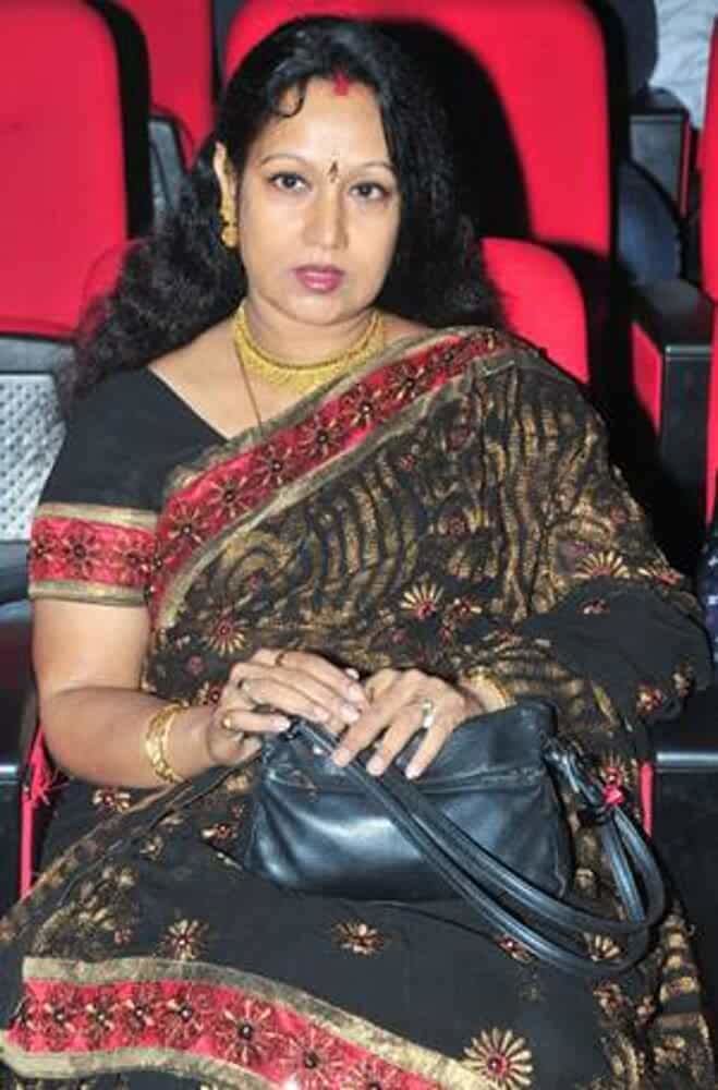 Prabha wearing earrings, a necklace, bracelets, rings, a black dress with a red floral design, and holding a black bag.