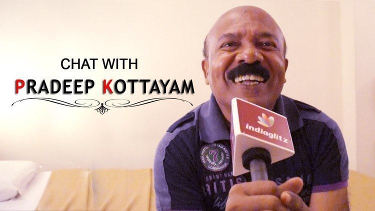 Pradeep Kottayam laughing with a bald head and a mustache while holding a microphone with indiaglitz logo in an interview called Chat with Pradeep Kottayam and wearing a purple shirt with prints