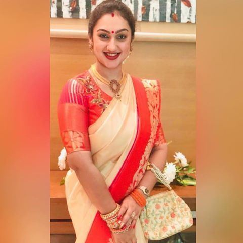 Preetha Vijayakumar smiling while carrying a bag and wearing a cream and orange dress and some pieces of jewelry
