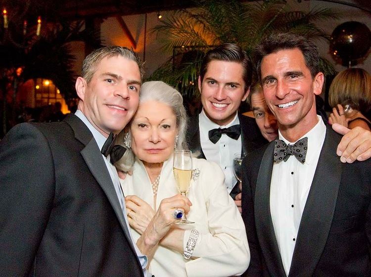 A man (left) is smiling, and has white hair, wearing powder blue long sleeves, and a black bow tie under a black suit. 2nd from left is Denise Hale being serious, has white hair, her right hand holding a glass of champagne and a diamond ring, her left hand is on her chest with a silver bracelet, wearing silver earrings, a necklace, and a white suit.  3rd from left is a man smiling, his right hand holding a glass of wine, has black hair, wearing white long sleeves with buttons and a black bow tie under a black suit. 4th from left is a man smiling, hand on the shoulder of the man (right) wearing a black suit. On (right) is a man smiling, has black hair, wearing white long sleeves with buttons, a gray bow tie with a  design of black flowers.