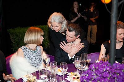 Anna Wintour (left) is sitting down on a violet chair and seriously looking at Gavin Newsom (right), who has a glass of water in front with different kinds of foods on the plate and a violet flower on the table, she has blonde short hair, wearing a necklace and a gray dress with a spiral design under a white coat. In the middle is Denise Hale seriously looking at Anna Wintour (left) while hugging Gavin Newsom (right) with her both hands, has white short hair, wearing a black dress. Gavin Newsom (right) is sitting down on a violet chair, seriously looking at Anna Wintour, both hands holding Denise Hale’s hands, he has a glass of water and wine on the table, wearing a white long sleeve, under a black coat.