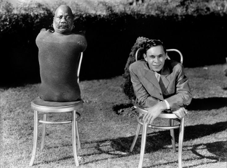 Johnny Eck smiling while sitting on the chair and Prince Randian with a serious face and mustache. Johnny Eck wearing a long sleeve under a coat while Prince Randian wearing a shirt