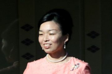 Princess Akiko of Mikasa is smiling while looking at something, wearing pearl earrings, a pearl necklace, and a pink dress with a pin badge on it.