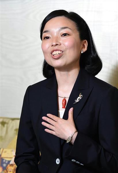 Princess Akiko of Mikasa is smiling while talking and her hand on her chest, with short black hair, wearing earrings, a red necklace, a watch, and a black blazer with a pin badge on it over a white top.