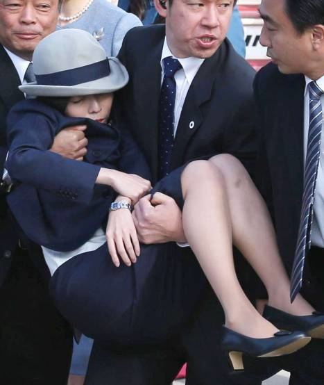 Princess Akiko of Mikasa collapsed and was carried by a security guard at Haneda Airport on December 6, 2013. Princess Akiko is wearing a gray and black hat, a watch on her left wrist, a black blazer over a white top, a black skirt, and black shoes while the security guards are wearing black suits, neckties, and white long sleeves, and black pants.