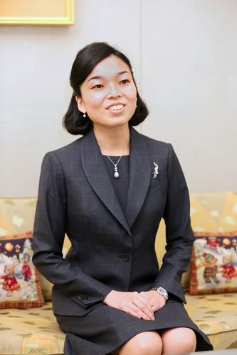 Princess Akiko of Mikasa is smiling while looking at something, sitting on a yellow couch and hands on her lap, with short black hair, wearing earrings, a necklace, a watch, a black skirt, and a black blazer with a pin badge on it over a black top.