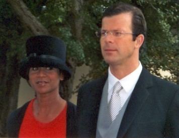 Princess Angela and Prince Maximilian of Liechtenstein with a tight-lipped smile while looking at something. Princess Angela is wearing a black hat and red dress under a black coat while Prince Maximilian is wearing eyeglasses and a white long sleeve under a light gray striped necktie, gray vest, and black coat