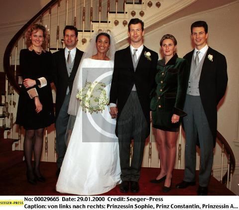Princess Sophie, Prince Constantin, Princess Angela, Prince Maximilian, Princess Marie, and Prince Alois (left to right) are smiling at the wedding ceremony. Princess Angela is holding a bouquet of flowers and wearing a white long-sleeve gown, white veil, and crown while Prince Maximilian is wearing dark gray pants and white long sleeve under a gray necktie, gray vest, and black coat