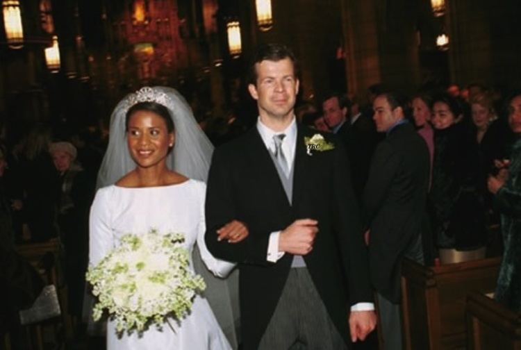 Princess Angela and Prince Maximilian of Liechtenstein are smiling during their wedding day. Princess Angela holding a bouquet of flowers  and wearing a white long-sleeve gown, white veil, and crown while Prince Maximilian is wearing dark gray pants and white long sleeve under a gray necktie, gray vest, and black coat