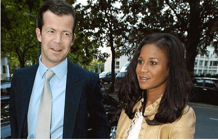 Prince Maximilian and Princess Angela of Liechtenstein are smiling while looking at something. Prince Maximilian is wearing a blue long sleeve under a gray necktie and black coat while Princess Angela is wearing a white blouse under a beige blazer