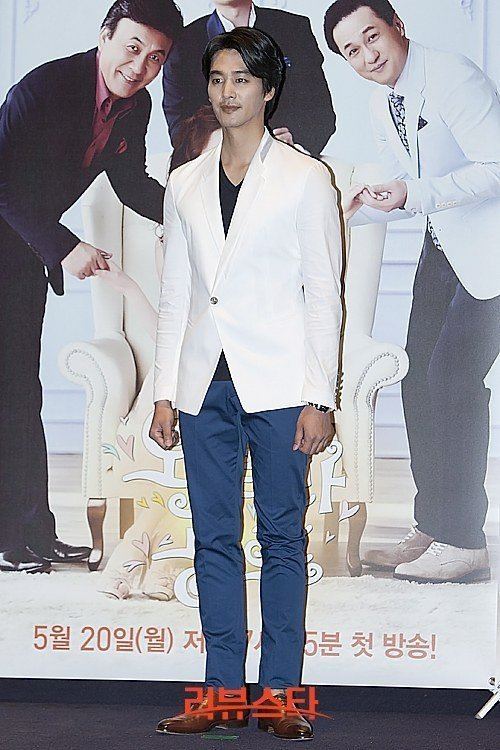 Oh Chang-Seok with a tight-lipped smile wearing a white coat, black inner shirt and blue pants