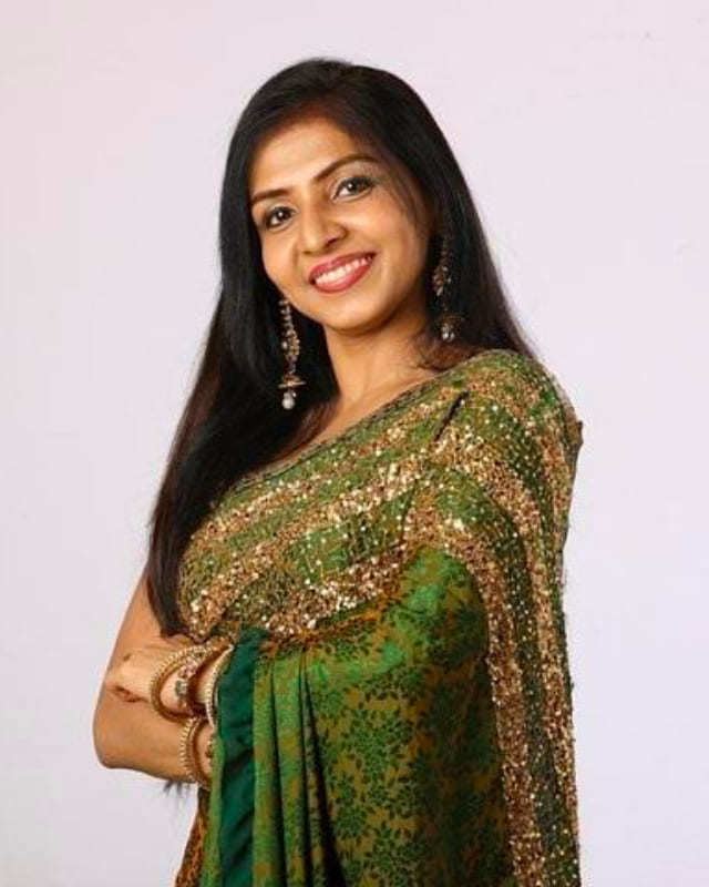 Priyadarshini is smiling, standing, arms crossed, has black hair wearing dangling earrings and gold bracelets, and a green printed with gold saree.