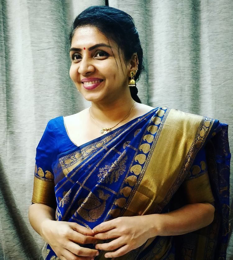 In a room with a large gray curtain, Priyadarshini is smiling, standing, with her hands together, has black hair, a bindi on her forehead, wearing gold dangling earrings, a gold necklace, and a blue top with gold printed saree.
