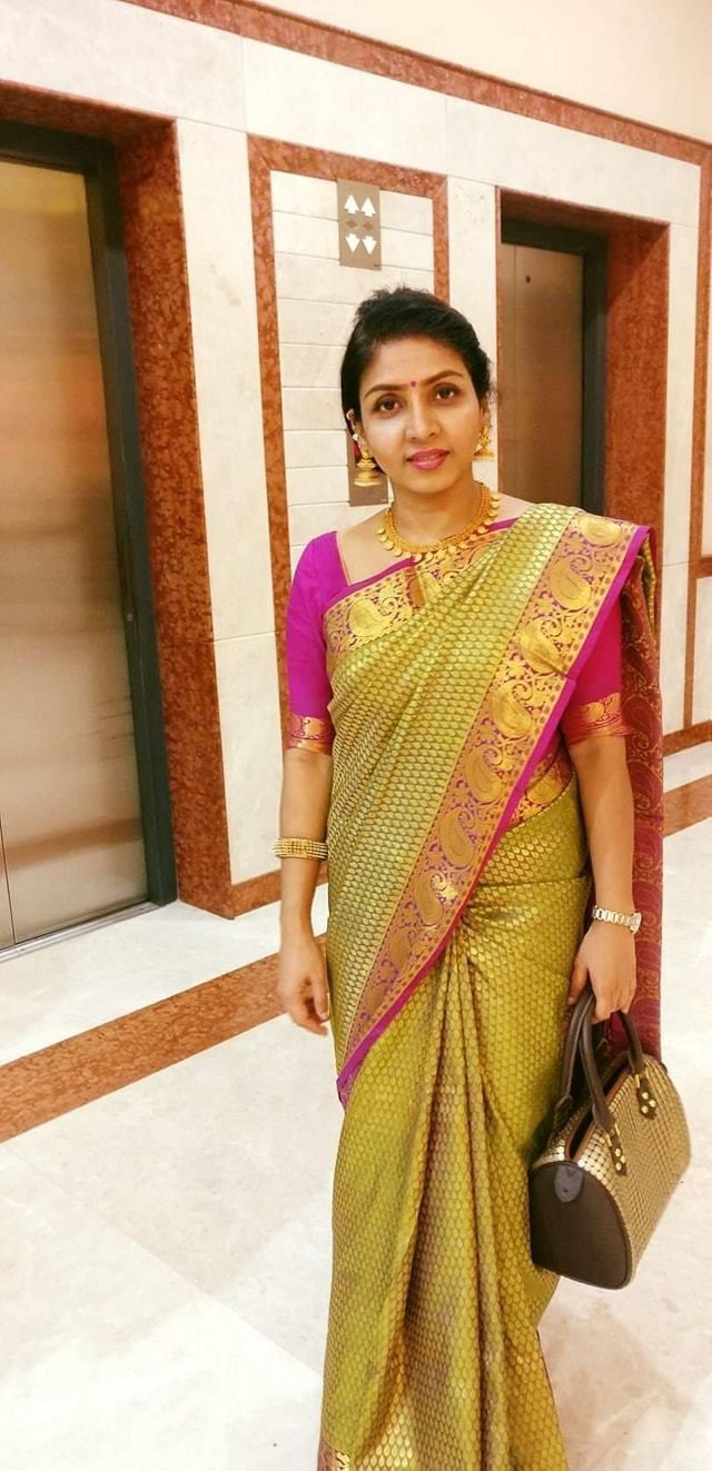 In a hotel hallway with elevators at the back, Priyadarshini is smiling, standing, with her hands down left hand holding a gold plated brown bag, has black hair, a bindi on her forehead, wearing gold dangling earrings, a gold necklace, gold bracelet, a silver watch and a purple top with gold printed saree.