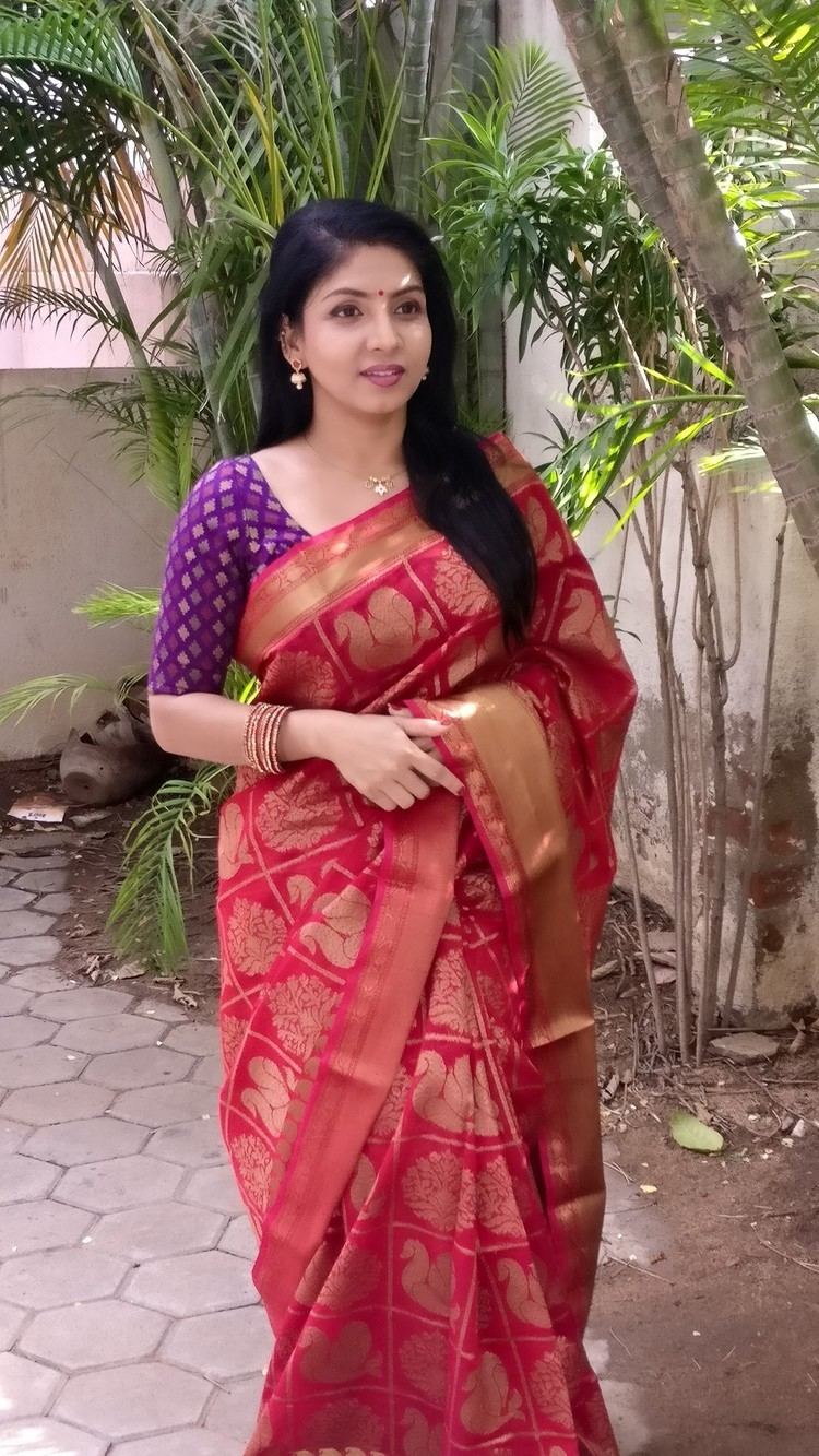 In a yard with white walls, has plants and trees beside a small pathway,  Priyadarshini is smiling, standing, holding her hands together, has black hair and a red bindi on her forehead, red fingernails, wearing a gold dangling earrings, a gold bracelet, a silver necklace and a purple top red printed saree.