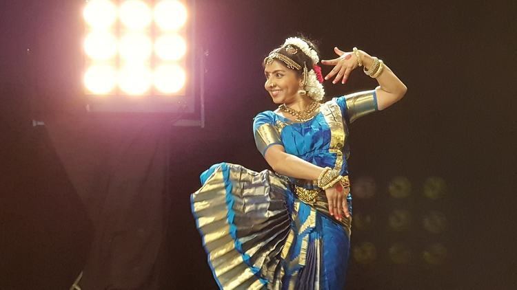 In a dark room with two large spotlights, Priyadarshini is smiling, dancing, looking at her right, standing with her left foot and right foot bent knee up, right hand down over her left waist, left hand up behind her head, has black hair with gold headband and a white flower tied up, a bindi on her forehead, wearing gold dangling earrings, a gold necklace, gold bracelets and a blue top with gold printed saree.