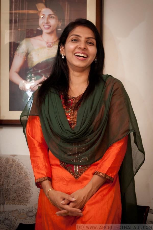 In a white room with Priyadarshini photo framed on the wall with a white sofa at the back, Priyadarshini is laughing, standing with her hands holding together, has black hair wearing pearl earrings, gold bracelets, an orange dress, and olive see through scarf.