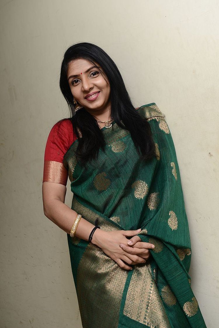 Priyadarshini is smiling, standing, with her hands together, has black hair, a bindi on her forehead, wearing earrings, a necklace with fan pendants, a gold and black bracelet and a red top with green saree.